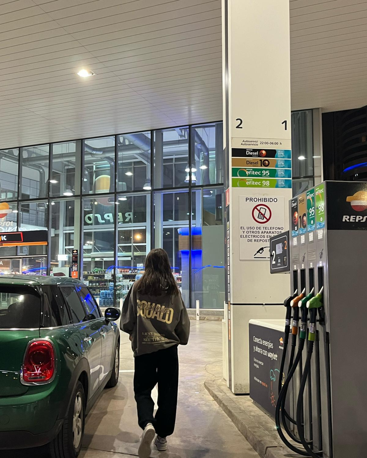 Mujer de espaldas con hoodie gris SQUALO con estampado dorado en una gasolinera de noche